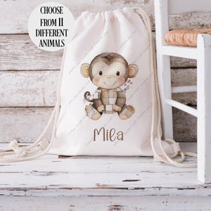 May include: A white drawstring bag with a brown monkey illustration and the name "Mila" printed on the front. The bag is made of natural fabric and has a simple design. Choose from 11 different animals.