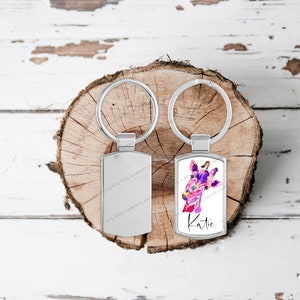 May include: Two silver keyrings with rectangular tags. One tag features a watercolour painting of a giraffe's head in shades of pink, purple and blue. The other tag is blank. The text "Katie" is printed on the tag with the giraffe.