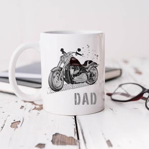 Op de afbeelding: Witte keramische mok met een zwart-witte illustratie van een motorfiets en het woord "DAD" gedrukt op de voorkant.