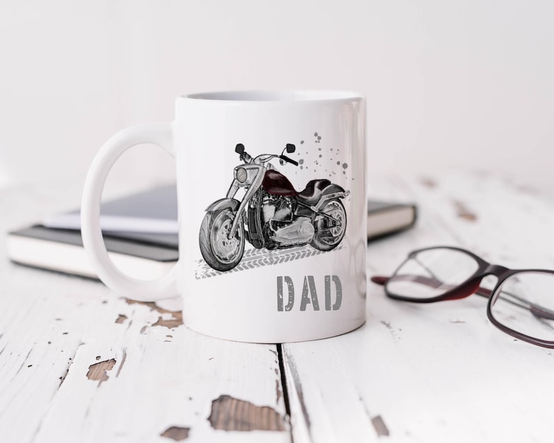 Op de afbeelding: Witte keramische mok met een zwart-witte illustratie van een motorfiets en het woord "DAD" gedrukt op de voorkant.