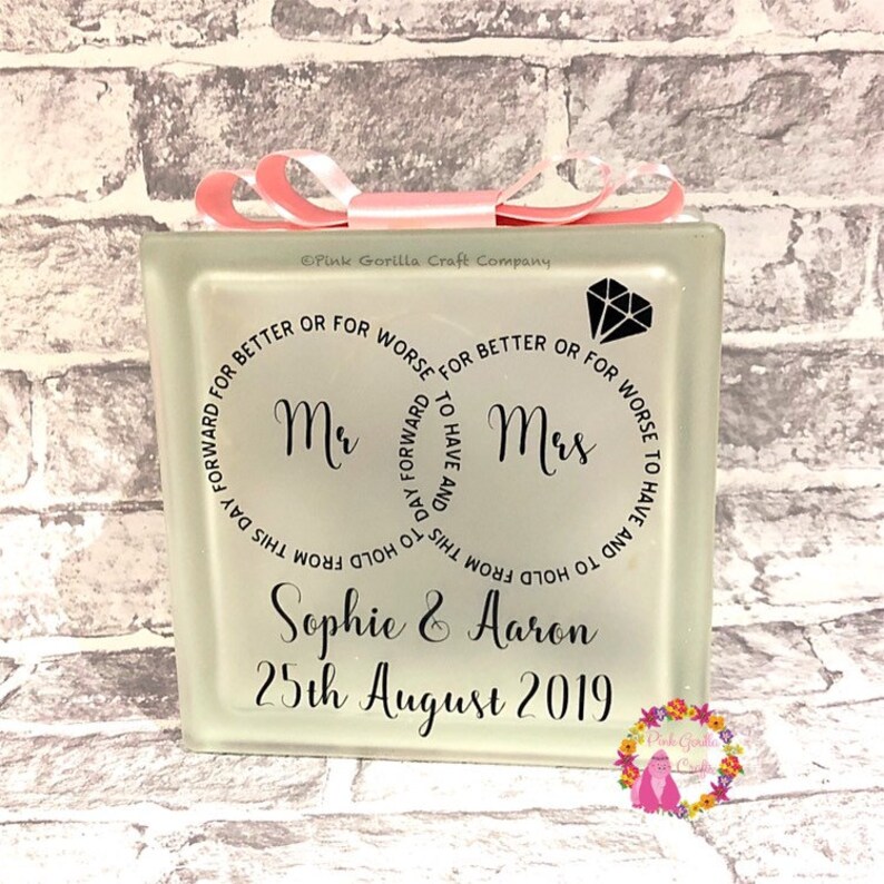 Op de afbeelding: Een witte glazen blok met een roze lint eromheen gebonden. De blok heeft zwarte tekst die luidt "Mr. & Mrs.  To have and to hold from this day forward for better or for worse." De namen "Sophie & Aaron" en de datum "25th August 2019" staan ook op de blok gedrukt.