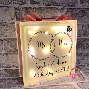 Op de afbeelding: Een wit, verlicht glazen blok met de woorden "Mr. & Mrs." en de tekst "Om te hebben en te houden vanaf deze dag, voor beter of voor slechter, om te hebben en te houden vanaf deze dag, voor beter of voor slechter." Het blok bevat ook de namen "Sophie & Aaron" en de datum "25 augustus 2019".