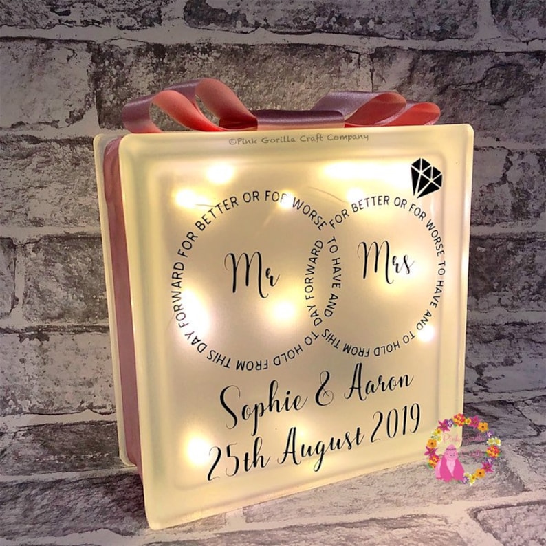 Op de afbeelding: Een wit, verlicht glazen blok met de woorden "Mr. & Mrs." en de tekst "Om te hebben en te houden vanaf deze dag, voor beter of voor slechter, om te hebben en te houden vanaf deze dag, voor beter of voor slechter." Het blok bevat ook de namen "Sophie & Aaron" en de datum "25 augustus 2019".