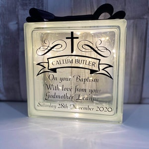 Op de afbeelding: Een heldere glazen blok met een kruis en de tekst "CALLUM BUTLER" op een banner. De tekst "On your Baptism With love from your Godmother Leanne Saturday 28th November 2020" is ook inbegrepen. De blok is verlicht met witte lichtsnoeren.