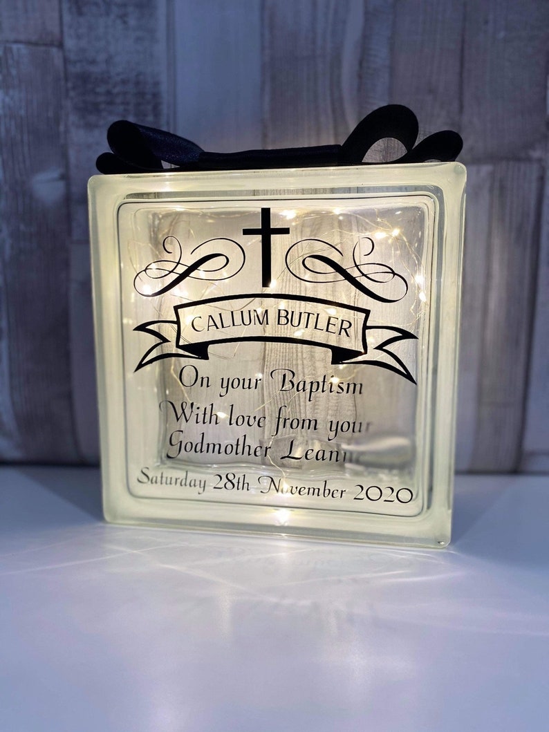 Op de afbeelding: Een heldere glazen blok met een kruis en de tekst "CALLUM BUTLER" op een banner. De tekst "On your Baptism With love from your Godmother Leanne Saturday 28th November 2020" is ook inbegrepen. De blok is verlicht met witte lichtsnoeren.
