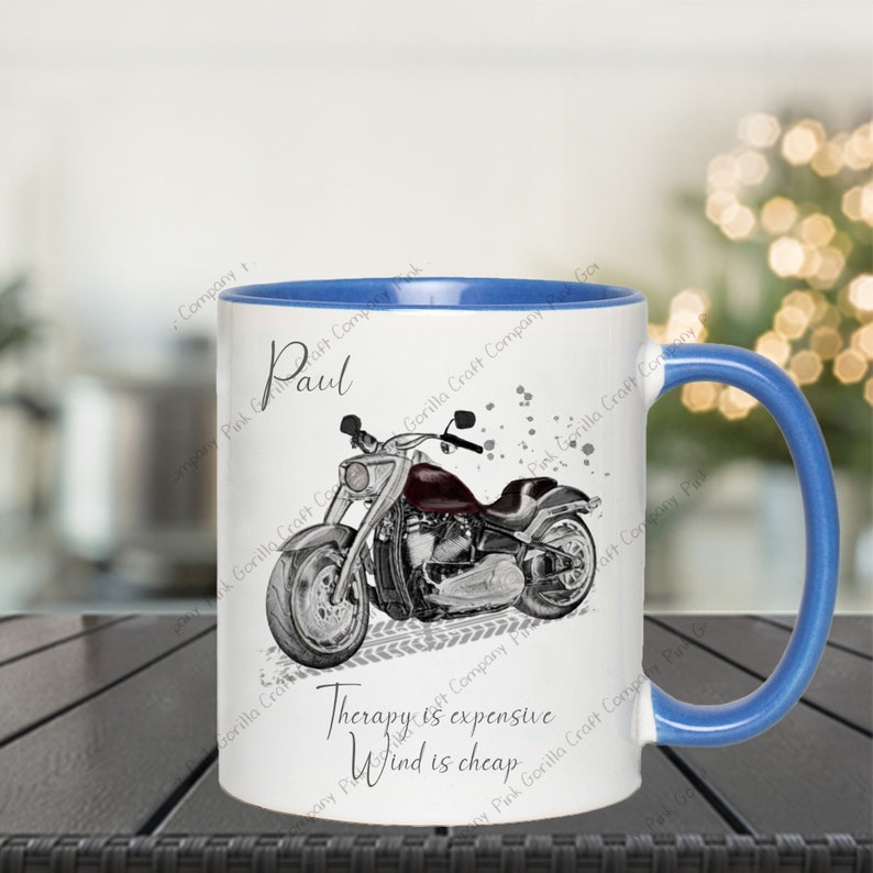 Op de afbeelding: Een witte keramische mok met een blauwe rand en handvat. De mok heeft een zwart-witte illustratie van een motorfiets met de tekst "Therapy is expensive, Wind is cheap". De naam "Paul" staat op de mok gedrukt.