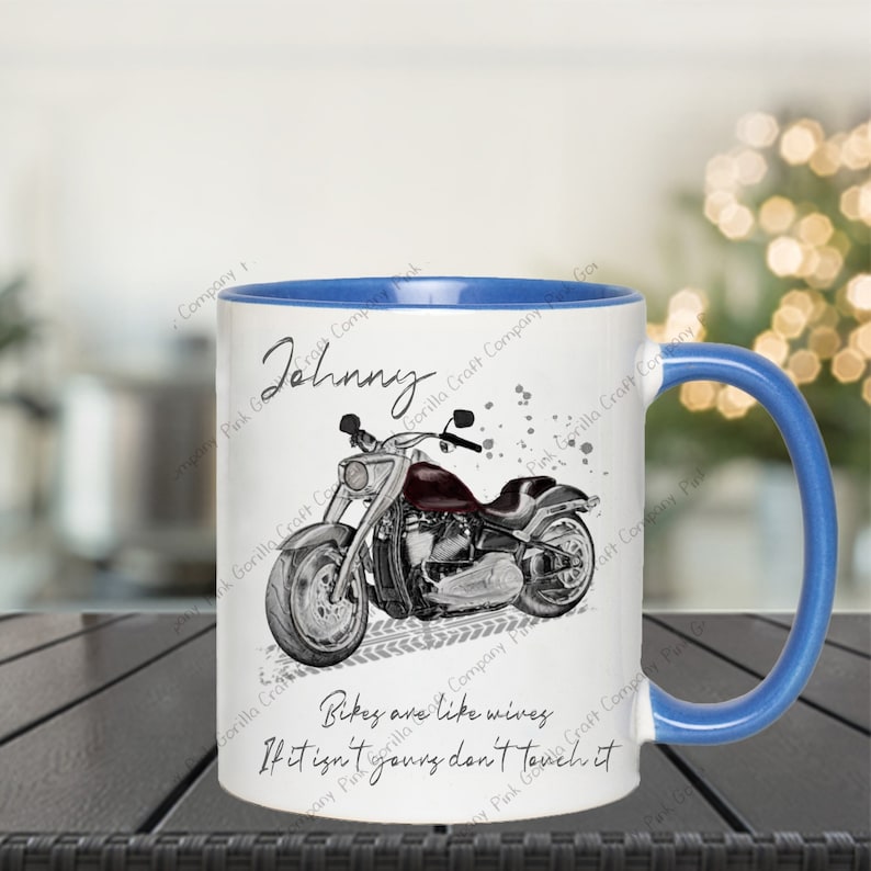 Op de afbeelding: Een witte keramische mok met een blauwe rand en handvat. De mok heeft een zwart-witte illustratie van een motorfiets met de tekst "Bikes are like wives. If it isn't yours don't touch it." en de naam "Johnny" bovenaan.