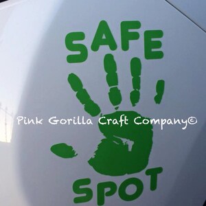 May include: A green handprint sticker with the words "SAFE SPOT" in green block letters. The sticker is on a white car surface. The text "Pink Gorilla Craft Company" is also visible.