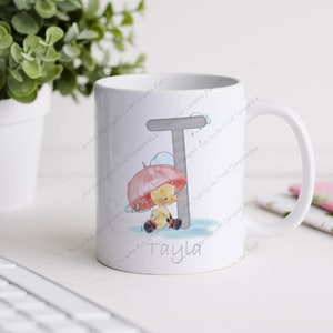May include: White ceramic mug featuring a grey letter T and a cartoon duckling holding a red umbrella. The name "Tayla" is written beneath the duckling.