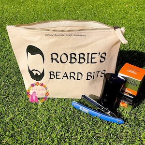 May include: A beige canvas zippered pouch with the text "ROBBIE'S BEARD BITS" printed in black. A black silhouette of a man with a beard is above the text. The pouch is sitting on a green grassy surface with a blue and silver straight razor, a black jar, and a green and orange box of beard care products.