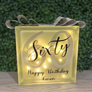 Op de afbeelding: Een verlicht glazen blok met het woord "Sixty" en de tekst "Happy Birthday Karen" in zwarte letters. Het blok is versierd met een zilveren lint.
