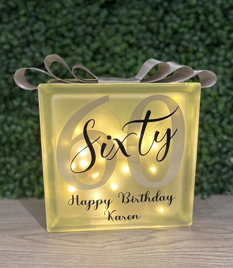 Op de afbeelding: Een verlicht glazen blok met het woord "Sixty" en de tekst "Happy Birthday Karen" in zwarte letters. Het blok is versierd met een zilveren lint.