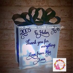 Op de afbeelding: Een wit, verlicht glazen blok met een zwart lint erop. Het blok heeft een blauwe inscriptie met de tekst "2005 To Helen 2016 Thank you for everything Love from Mia Xxxx".