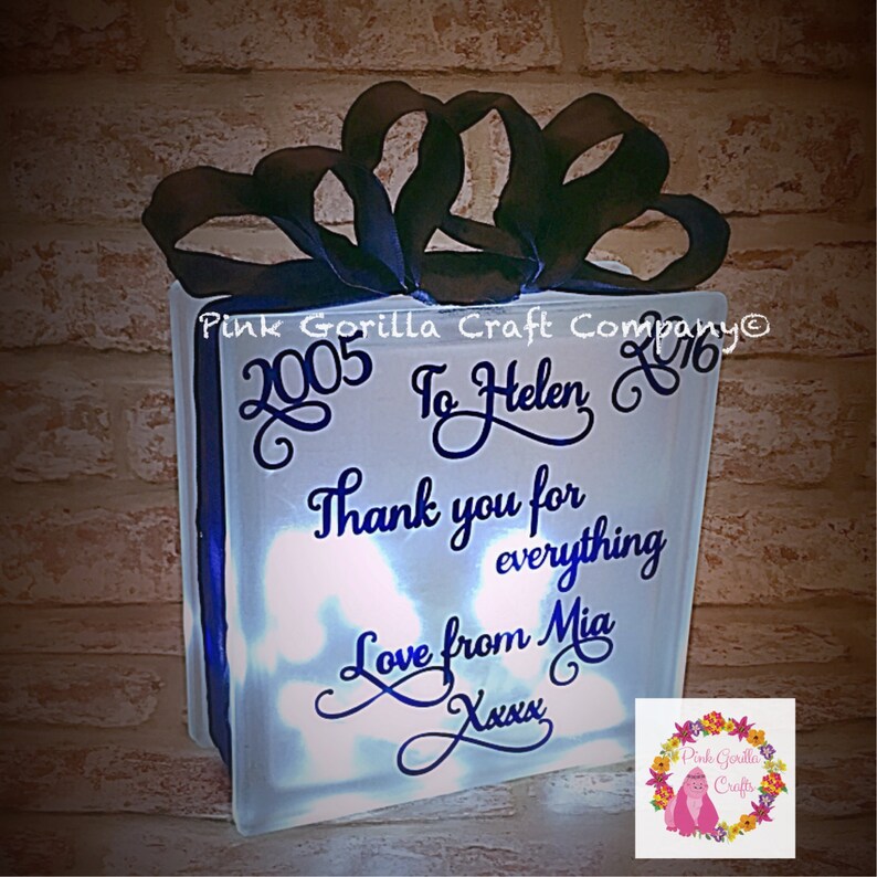 Op de afbeelding: Een wit, verlicht glazen blok met een zwart lint erop. Het blok heeft een blauwe inscriptie met de tekst "2005 To Helen 2016 Thank you for everything Love from Mia Xxxx".