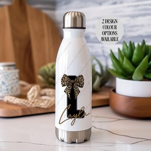 May include: A white insulated water bottle with a silver lid and base. The bottle features a black letter "L" with a leopard print bow and the name "Kayla" in gold script. The text "2 DESIGN COLOUR OPTIONS AVAILABLE" is also visible.