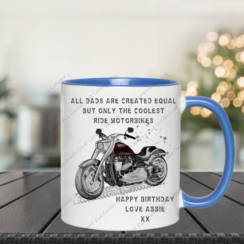 Op de afbeelding: Een witte keramische mok met een blauwe rand en handvat. De mok heeft een zwart-witte illustratie van een motorfiets met de tekst "Alle vaders zijn gelijk geschapen, maar alleen de coolste rijden motorfiets". De mok heeft ook de tekst "Gefeliciteerd met je verjaardag Lieve Abbie XX".