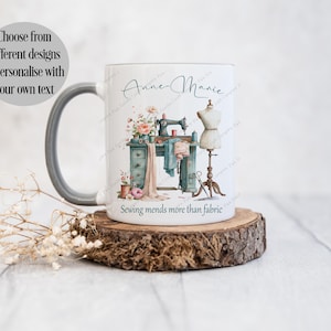 May include: A white ceramic mug with a grey handle featuring a printed illustration of a vintage sewing machine on a desk, a dress form, and floral accents. The text reads "Anne-Marie" and "Sewing mends more than fabric" in black script and print.