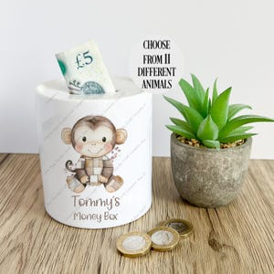May include: White ceramic money box featuring a cartoon monkey wearing a plaid shirt. The box is labelled "Tommy's Money Box". A five pound note is partially inserted into the slot on the top of the box. The text "CHOOSE FROM 11 DIFFERENT ANIMALS" is visible in the background.
