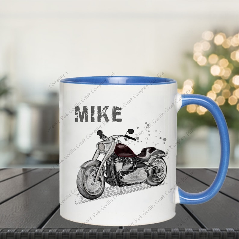 Op de afbeelding: Een witte keramische mok met een blauwe rand en handvat. De mok heeft een zwart-witte illustratie van een motorfiets met de naam "MIKE" in zwart op de voorkant.