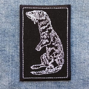 Op de afbeelding: Een rechthoekige zwarte patch met een witte geborduurde illustratie van een staande fret. De fret is gedetailleerd en gestileerd afgebeeld, met zichtbare vacht en gelaatstrekken. De patch heeft een witte rand en staat op een blauwe denim achtergrond.