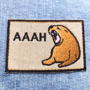 Op de afbeelding: Geborduurde patch met een schreeuwende cartoonbeer, met het woord "AAAH" erboven. De patch heeft een beige achtergrond en een zwarte rand. De patch is geplaatst tegen een blauwe denim achtergrond.