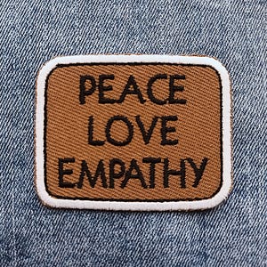 May include: A rectangular patch with the words "PEACE LOVE EMPATHY" in black lettering. The patch has a brown background with a white border, set against a blue denim fabric.