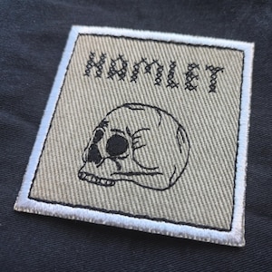 Könnte beinhalten: Quadratischer Aufnäher mit dem Wort "HAMLET" in Schwarz über einem schwarzen Totenkopf-Design gestickt. Der Aufnäher hat einen beigefarbenen Hintergrund mit weißem Rand, vor dunklem Stoff.