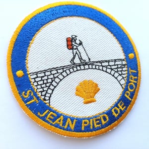 Könnte beinhalten: Runder, gestickter Aufnäher mit blau-gelbem Rand. Er zeigt einen Pilger auf einer Brücke, eine gelbe Jakobsmuschel und den Text "ST JEAN PIED DE PORT".