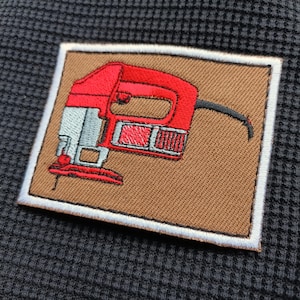 May include: Embroidered patch featuring a red jigsaw on a brown background with a white border. The patch is rectangular and is set against a dark gray textured fabric.