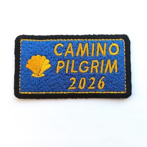 Op de afbeelding: Geborduurde patch met de woorden "CAMINO PILGRIM 2026" in geel op een blauwe achtergrond. Links staat een gele sint-jakobsschelp. De patch heeft een zwarte rand.