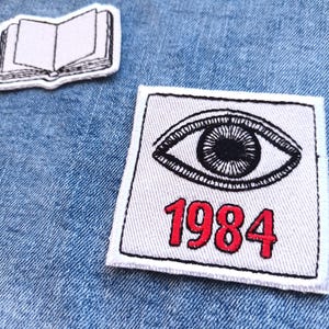 May include: Two embroidered patches on denim. One patch shows an open book illustration. The other patch has an eye design above the red text "1984". Both patches have a white background and black border.