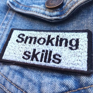 May include: A rectangular patch with the words "Smoking skills" embroidered in black on a white background. The patch has a black border and is sewn onto a blue denim jacket.
