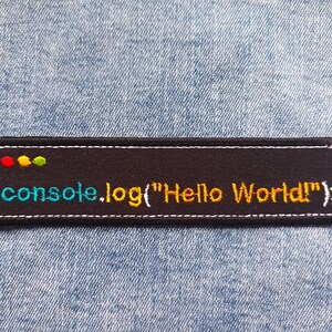 Puede incluir: Un parche bordado negro con el texto "console.log("Hello World!");" en verde azulado y dorado. El parche tiene un borde blanco y puntos coloridos en la parte superior, sobre un fondo de mezclilla. Un accesorio divertido para programadores.