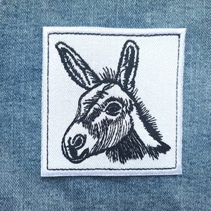 Puede incluir: Un parche cuadrado bordado en blanco con una detallada ilustración en blanco y negro de la cabeza de un burro. El parche tiene un borde cosido blanco y está sobre un fondo de mezclilla azul.