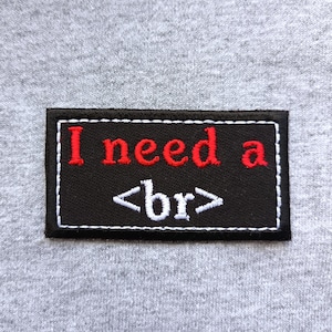 Puede incluir: Un parche rectangular negro con costuras blancas y las palabras "I need a <br>" bordadas en rojo y blanco. El parche está sobre un fondo de tela gris.