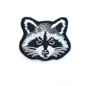 May include: Embroidered patch featuring a raccoon face. The patch has a black background with white embroidered details, including the eyes, nose, and fur. The raccoon's face is centered, with a detailed and expressive design. The patch is likely for clothing or accessories.