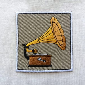 Könnte beinhalten: Gestickter Aufnäher mit einem Vintage-Grammophon. Das Grammophon hat einen großen, gelben Trichter und einen braunen Sockel. Der Aufnäher ist quadratisch mit grauem Hintergrund und weißem Rand. Das Design ist detailliert und maschinengestickt.