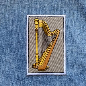 Op de afbeelding: Geborduurde patch met een gouden harp op een rechthoekige, grijsbeige achtergrond met een witte rand. De harp heeft snaren en een gebogen klankkast. De patch is geplaatst tegen een blauwe denim achtergrond.