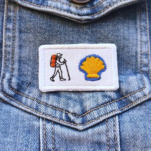 Può includere: Toppa ricamata raffigurante un pellegrino con zaino e bastone da passeggio, e una conchiglia gialla su sfondo bianco. La toppa è rettangolare e cucita su una tasca di giacca di jeans.