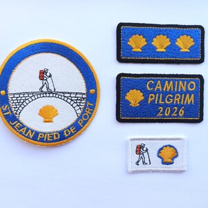 May include: Embroidered patches featuring a pilgrim with a backpack, a bridge, and a scallop shell. One patch reads "ST JEAN PIED DE PORT". Other patches have scallop shells and the text "CAMINO PILGRIM 2026".