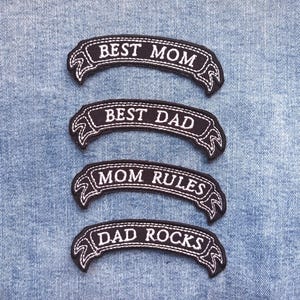 May include: Four black and white embroidered patches with the phrases "BEST MOM", "BEST DAD", "MOM RULES", and "DAD ROCKS". The patches are in a banner shape with decorative edges, and are sewn onto a light blue denim background.