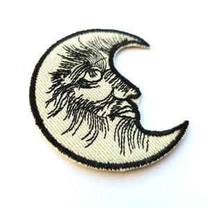 May include: Embroidered crescent moon patch with a face, featuring black thread on a cream-colored background. The patch has a black border and is likely for clothing or accessories.