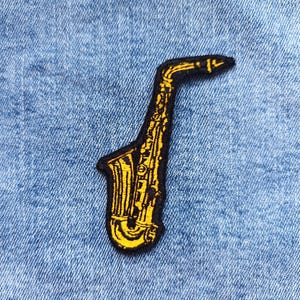 Pode incluir: Um patch de saxofone amarelo com borda preta, sobre um fundo de ganga azul. O patch apresenta linhas e curvas detalhadas, retratando a forma do instrumento. Ideal para amantes da música e músicos.