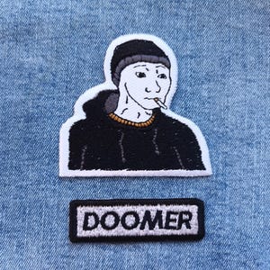 Op de afbeelding: Geborduurde patchset met een meme-personage in een zwarte hoodie en beanie, die een sigaret rookt. Hieronder een rechthoekige patch met het woord "DOOMER" in witte letters op een zwarte achtergrond. De patches liggen op een blauwe denim achtergrond.
