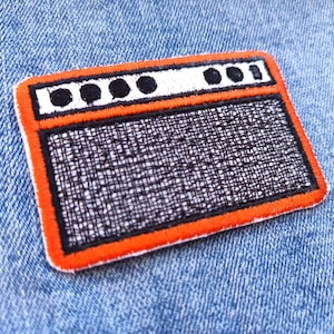Peut inclure: Un écusson brodé représentant un amplificateur orange et noir vintage. L'écusson a un dessus blanc avec quatre cercles noirs et une zone de haut-parleur en maille noire. L'écusson est sur un fond en denim bleu.