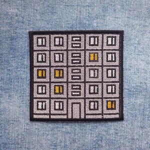 Könnte beinhalten: Quadratischer Aufnäher mit einem grauen Gebäudefassaden-Design mit schwarzen Umrissen und einem schwarzen Rand. Die Fenster sind weiß und grau, mit ein paar gelben Fenstern. Der Aufnäher befindet sich auf einem blauen Jeans-Hintergrund.