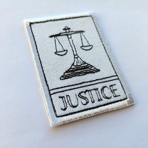 Può includere: Toppa ricamata con un disegno in bianco e nero della bilancia della giustizia. La parola "JUSTICE" è ricamata in nero sotto la bilancia. La toppa ha uno sfondo bianco e un bordo argentato.