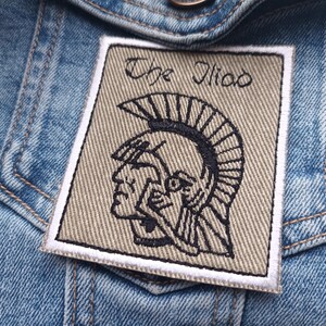 May include: A square patch featuring the text "The Iliad" and a black and white illustration of a warrior's helmet. The beige patch with a white border is sewn onto a blue denim jacket.