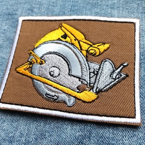May include: Embroidered patch featuring a circular saw design. The patch has a brown background with a white border. The saw is silver and yellow, with detailed stitching. The patch is square and likely for application on clothing or accessories.