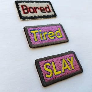 May include: Three embroidered patches with text. The top patch reads "Bored" in red thread on a white background with a black border. The middle patch reads "Tired" in yellow thread on a pink background with a gray border. The bottom patch reads "SLAY" in yellow thread on a pink background with a gray border.